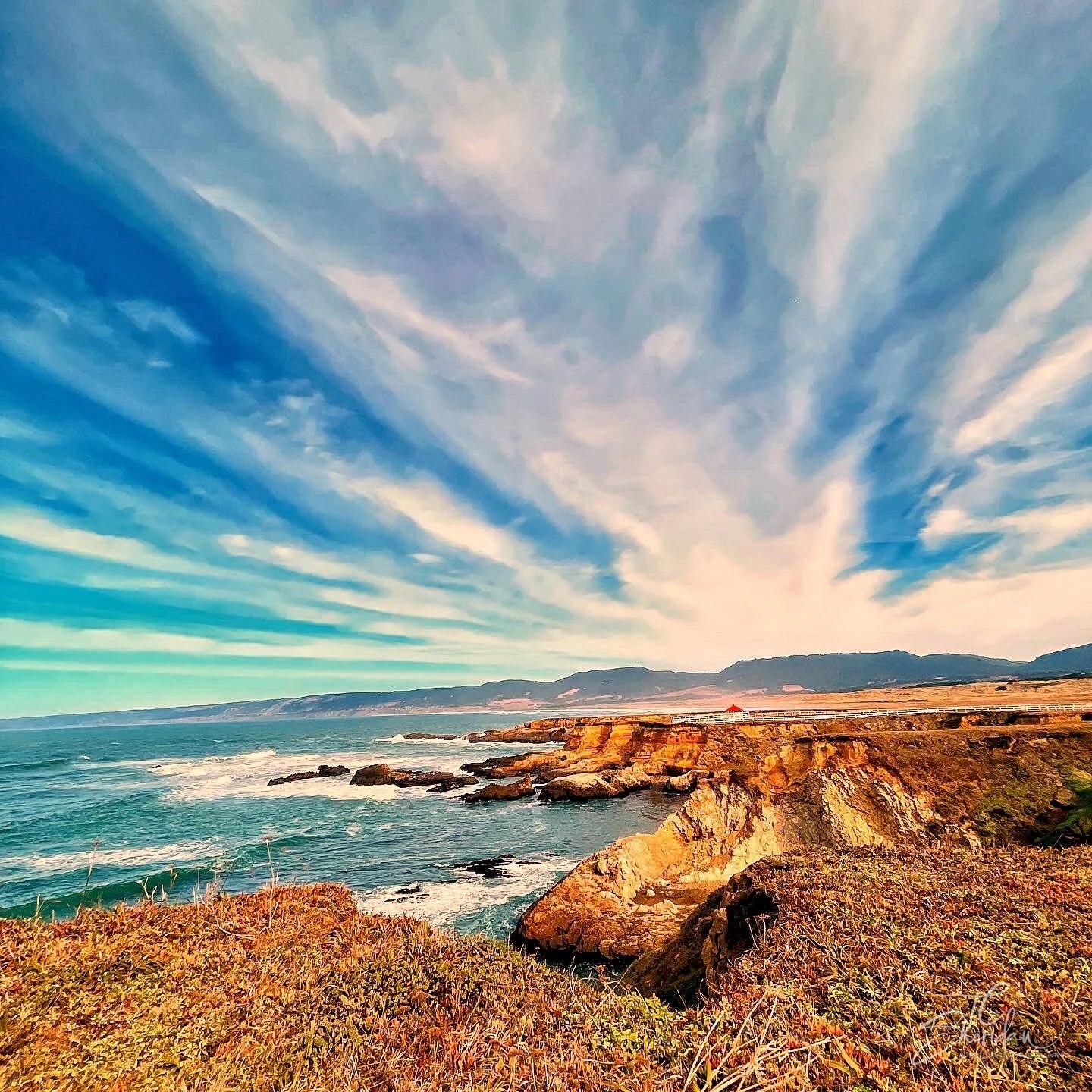 Sweeping clouds over turquoise water and a sunlit coastal headland, photographed by TC Sheridan